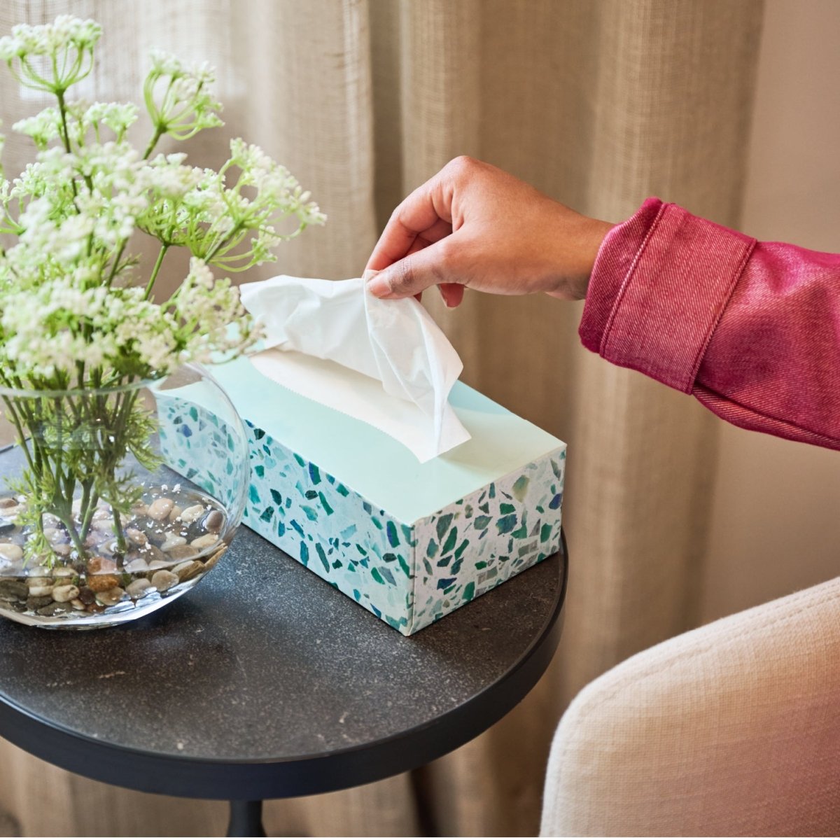 Hand reaching for a tissue from a teal terrazzo box on a table with flowers.